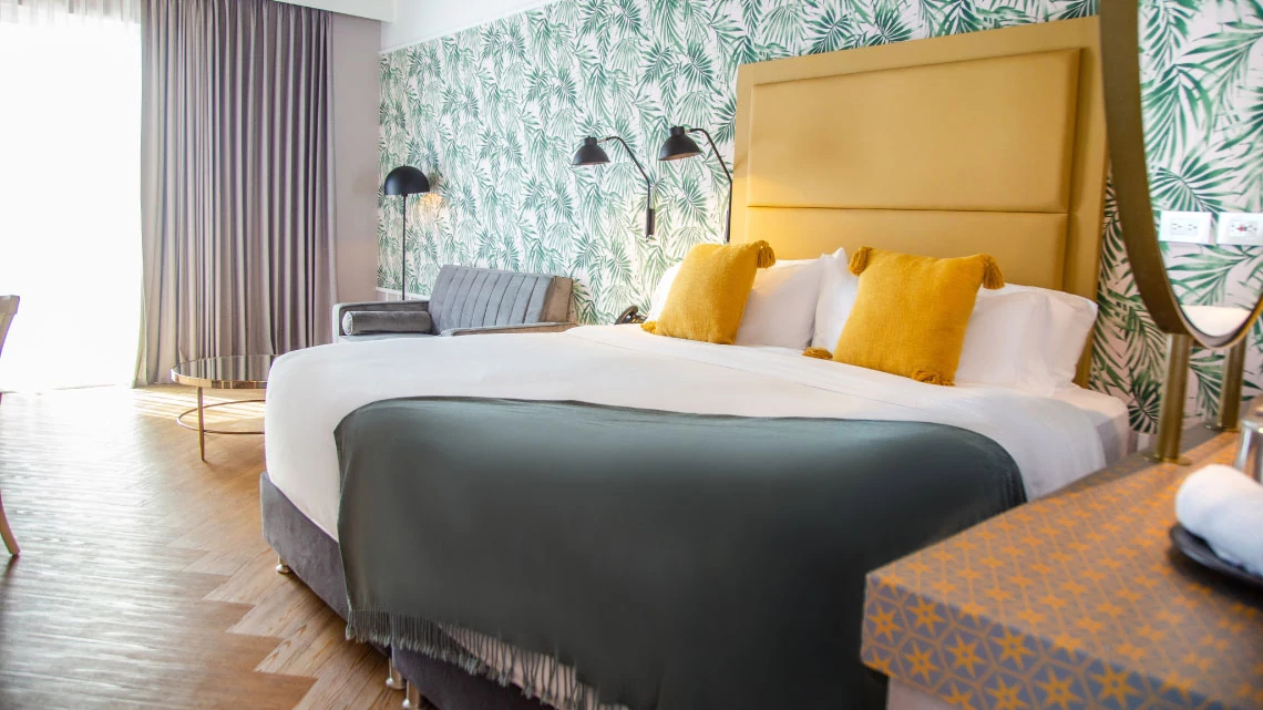 green, white, yellow linens on bed in hotel room with gray couch and colorful wallpaper