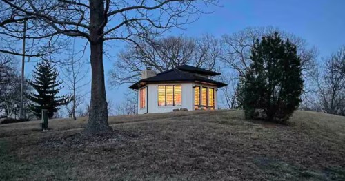 Distant, twilight view of the small, illuminated white octagonal house perched on a grassy hill surrounded by trees.