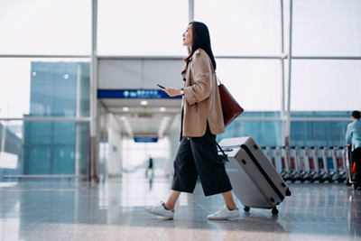 شابة آسيوية تحمل حقيبة سفر وتمسك بهاتف ذكي في يدها، تسير في صالة المطار