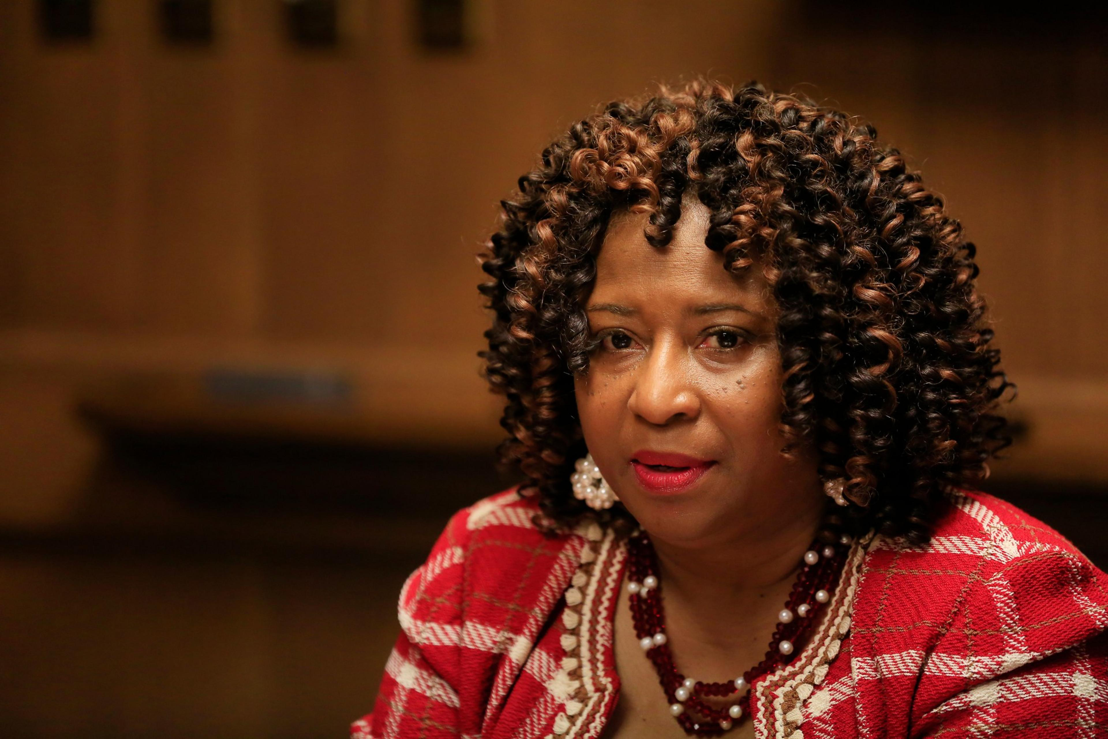 A woman with curly hair wears a red and white patterned jacket, pearl earrings, and a matching necklace, against a blurred brown background.