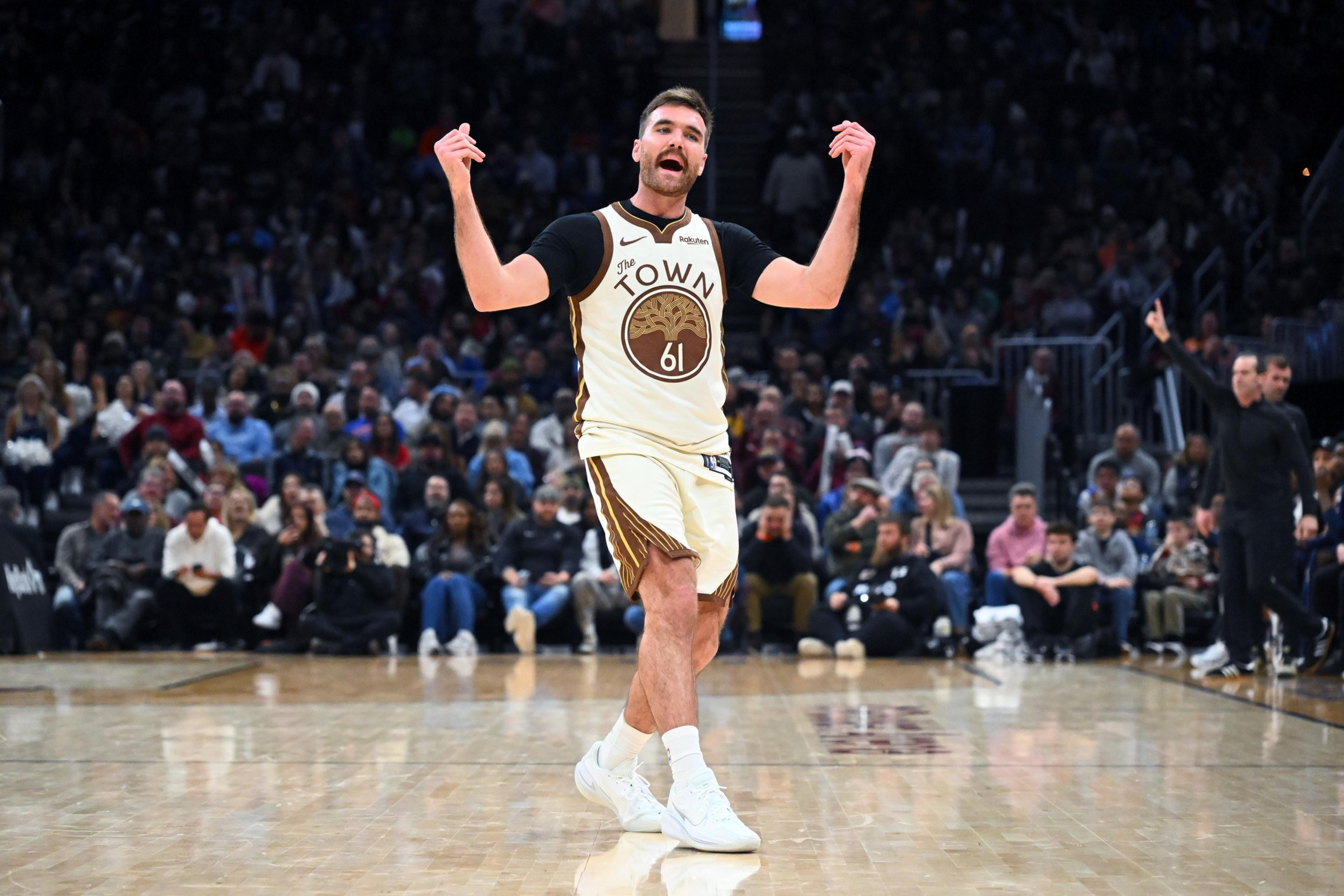 A basketball player wearing a white and brown "The Town" jersey with number 61 gestures with both hands on the court, while a crowd watches behind him.