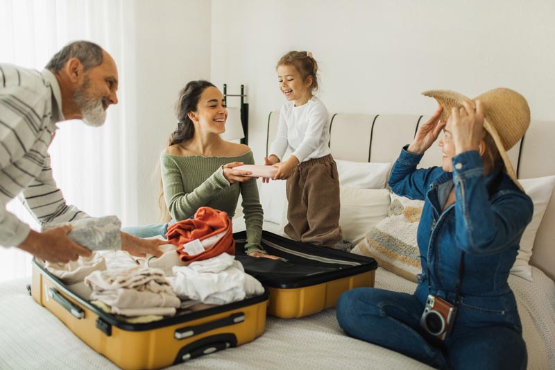 AARP Smart Guides Three adults and one kid sitting on a bed with an open suitcase full of clothes