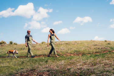 Two hikers and a dog; the hikers are wearing Allover Apparel overalls