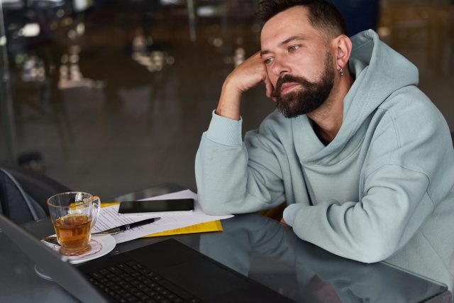 Pensive bearded man in casual clothes in cafe using his laptop