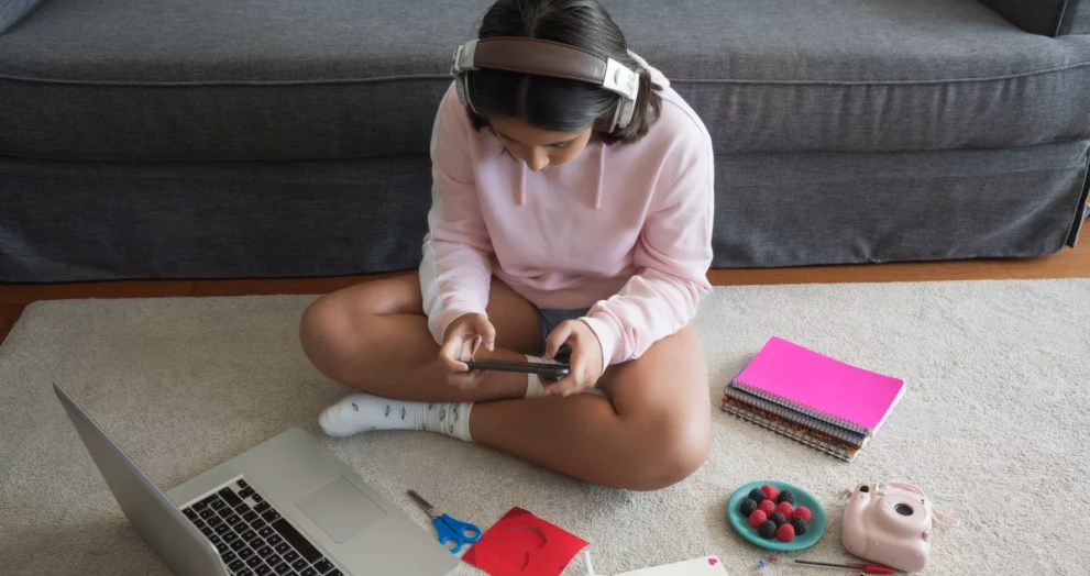 girl with headphones and a pink sweatshirt playing on her phone while her laptop, scissors, notebook, ad pens are by her