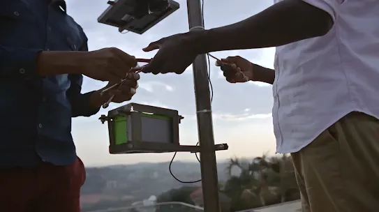 Two individuals shown from the chest down work with wires and a small box attached to a pole with a solar panel against a dusky sky background.